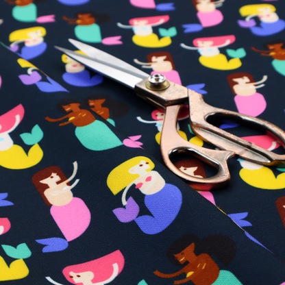 A fun and vibrant children's fabric on a deep midnight-navy background. The pattern features a diverse cast of illustrated mermaids with various skin tones, hair colours (pink, blonde, brown), and colourful tails in shades of mint, yellow, pink, and purple, arranged in a cheerful repeating grid.