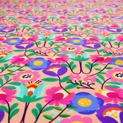 A vibrant and energetic floral fabric on a soft peach-blush background. The design features a symmetrical folk-art arrangement of stylized flowers in neon pink, electric blue, violet, and turquoise, connected by emerald green leaves and delicate black stems in a dense, mirrored repeat.