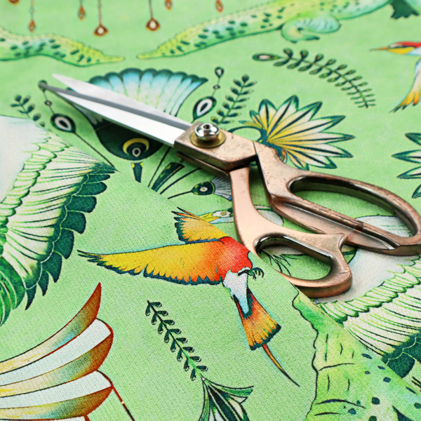 Ornate upholstery fabric with a mirrored botanical and wildlife design. Features white herons with outspread wings, crocodiles, hummingbirds, and butterflies surrounding stylized teal and gold fan motifs on a soft lime-green base.