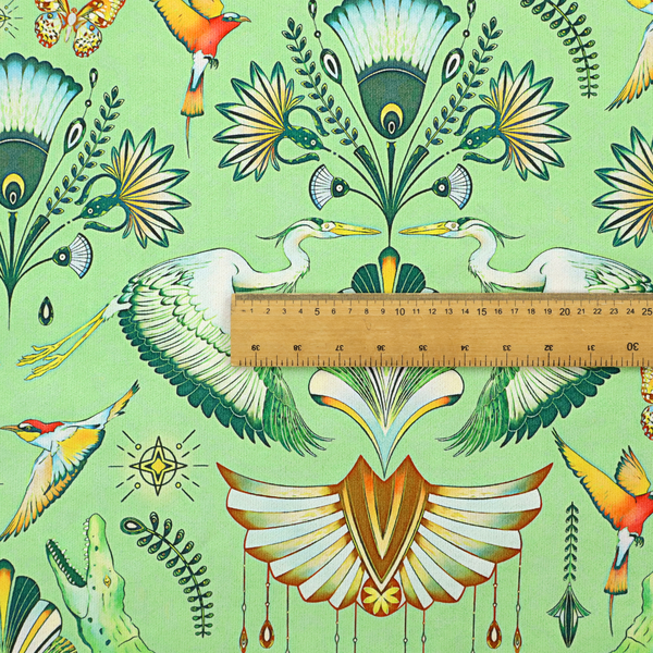 Ornate upholstery fabric with a mirrored botanical and wildlife design. Features white herons with outspread wings, crocodiles, hummingbirds, and butterflies surrounding stylized teal and gold fan motifs on a soft lime-green base.