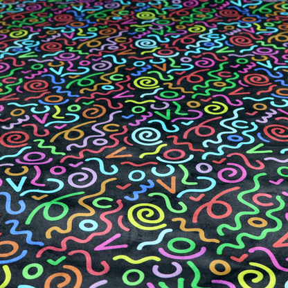A black fabric print featuring a retro, 90s-inspired pattern of colourful, hand-drawn-style doodles. The design includes vibrant neon swirls, squiggles, and V-shapes in shades of pink, yellow, blue, orange, and green, creating a nostalgic, high-energy, and playful aesthetic.