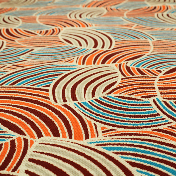 This fabric features a geometric, fan-like pattern composed of overlapping semi-circular arcs. The design creates a dynamic sense of movement through a vibrant colour palette of burnt orange, deep brown, bright turquoise, and cream.