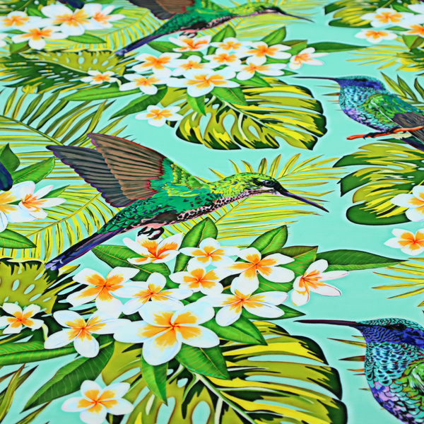 This fabric features a vibrant, tropical pattern set against a bright turquoise background. The design showcases hummingbirds in various poses—some perched and others in flight—surrounded by clusters of white plumeria flowers and lush, layered palm and monstera leaves in shades of green and yellow.