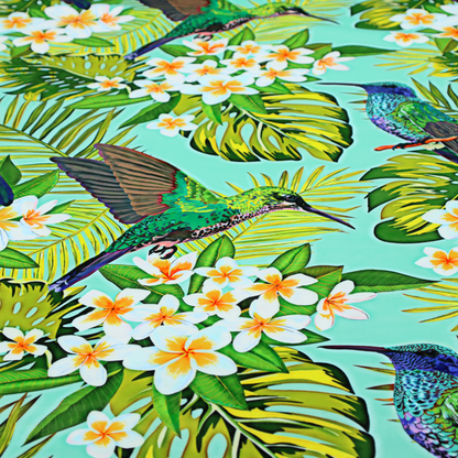 This fabric features a vibrant, tropical pattern set against a bright turquoise background. The design showcases hummingbirds in various poses—some perched and others in flight—surrounded by clusters of white plumeria flowers and lush, layered palm and monstera leaves in shades of green and yellow.