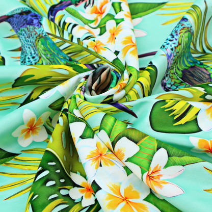 This fabric features a vibrant, tropical pattern set against a bright turquoise background. The design showcases hummingbirds in various poses—some perched and others in flight—surrounded by clusters of white plumeria flowers and lush, layered palm and monstera leaves in shades of green and yellow.
