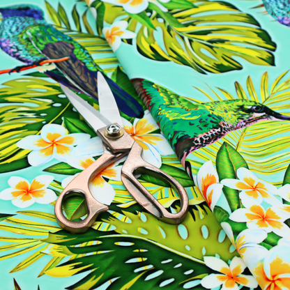 This fabric features a vibrant, tropical pattern set against a bright turquoise background. The design showcases hummingbirds in various poses—some perched and others in flight—surrounded by clusters of white plumeria flowers and lush, layered palm and monstera leaves in shades of green and yellow.