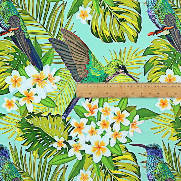This fabric features a vibrant, tropical pattern set against a bright turquoise background. The design showcases hummingbirds in various poses—some perched and others in flight—surrounded by clusters of white plumeria flowers and lush, layered palm and monstera leaves in shades of green and yellow.