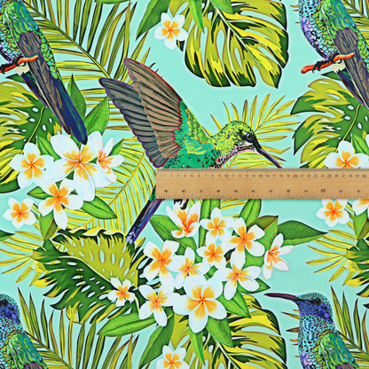 This fabric features a vibrant, tropical pattern set against a bright turquoise background. The design showcases hummingbirds in various poses—some perched and others in flight—surrounded by clusters of white plumeria flowers and lush, layered palm and monstera leaves in shades of green and yellow.
