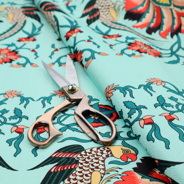 This fabric features an intricate, symmetrical folk-art inspired pattern set against a soft, light turquoise background. The design showcases stylized birds facing toward central, ornate floral bursts, rendered in a bold colour palette of vibrant red, deep orange, teal, and charcoal.