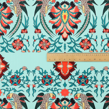 This fabric features an intricate, symmetrical folk-art inspired pattern set against a soft, light turquoise background. The design showcases stylized birds facing toward central, ornate floral bursts, rendered in a bold colour palette of vibrant red, deep orange, teal, and charcoal.