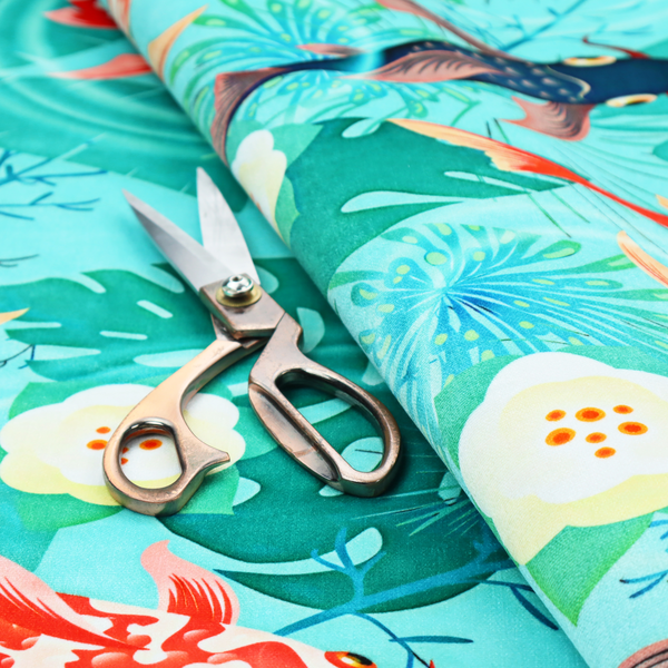 Patterned fabric featuring vibrant orange, white, and dark blue koi fish swimming among lush tropical monstera leaves, water ripples, and white flowers on a light teal background.