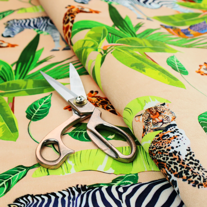 Patterned fabric featuring a vibrant safari scene with giraffes, zebras, leopards, and snakes nestled among lush tropical palm leaves on a soft beige background.
