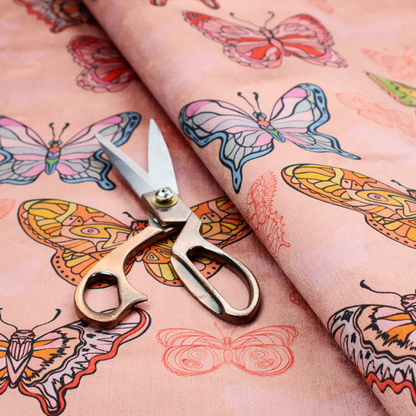 Patterned fabric featuring a whimsical, repeating design of colourful butterflies in various patterns and shades—including orange, teal, pink, and yellow—scattered among faint, outline-style butterfly silhouettes on a soft, dusty pink background.