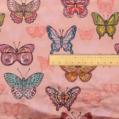 Patterned fabric featuring a whimsical, repeating design of colourful butterflies in various patterns and shades—including orange, teal, pink, and yellow—scattered among faint, outline-style butterfly silhouettes on a soft, dusty pink background.