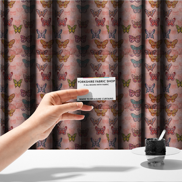 The fabric used to create these made-to-measure curtains is a patterned fabric featuring a whimsical, repeating design of colourful butterflies in various patterns and shades—including orange, teal, pink, and yellow—scattered among faint, outline-style butterfly silhouettes on a soft, dusty pink background.