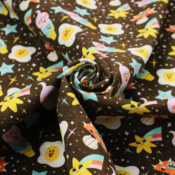 A whimsical, repeating fabric pattern featuring cartoon-style fried eggs, happy ice cream cones, shooting stars, and colourful stars on a dark chocolate brown background.