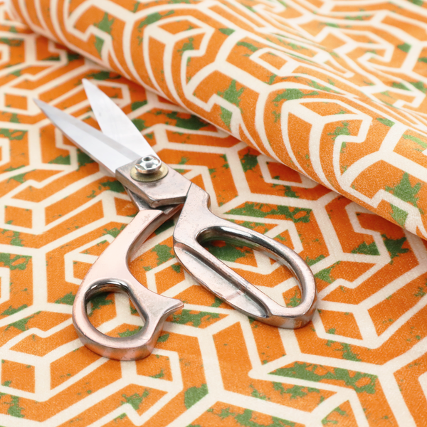 An orange and white textile fabric featuring a repeating, interlocking geometric pattern. The design consists of white outlines that form three-dimensional, maze-like structures or isometric blocks against a vibrant orange background. A textured, green moss-like speckled effect is scattered across the orange spaces, adding an organic contrast to the sharp, structured geometric lines.