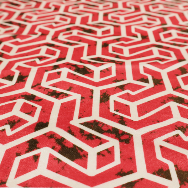 A red and off-white textile fabric featuring a repeating, interlocking geometric pattern. The design consists of off-white outlines forming three-dimensional, isometric maze-like structures against a vibrant coral red background. Distressed, dark brown speckled textures are subtly scattered throughout the red spaces, adding a weathered, organic touch to the structured, geometric design.