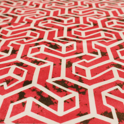 A red and off-white textile fabric featuring a repeating, interlocking geometric pattern. The design consists of off-white outlines forming three-dimensional, isometric maze-like structures against a vibrant coral red background. Distressed, dark brown speckled textures are subtly scattered throughout the red spaces, adding a weathered, organic touch to the structured, geometric design.