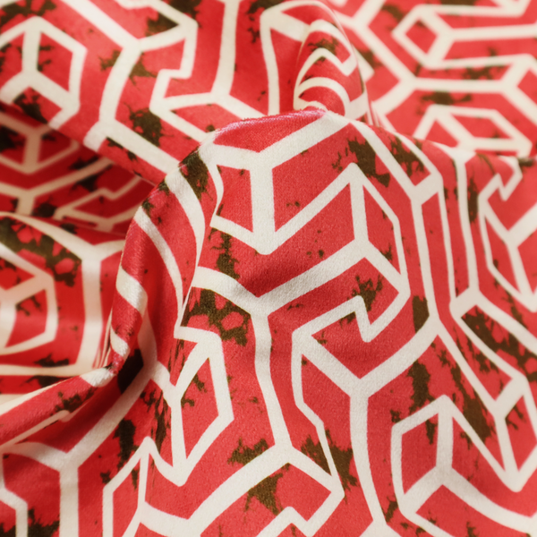 A red and off-white textile fabric featuring a repeating, interlocking geometric pattern. The design consists of off-white outlines forming three-dimensional, isometric maze-like structures against a vibrant coral red background. Distressed, dark brown speckled textures are subtly scattered throughout the red spaces, adding a weathered, organic touch to the structured, geometric design.