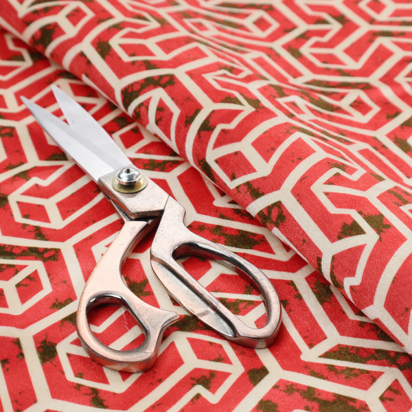 A red and off-white textile fabric featuring a repeating, interlocking geometric pattern. The design consists of off-white outlines forming three-dimensional, isometric maze-like structures against a vibrant coral red background. Distressed, dark brown speckled textures are subtly scattered throughout the red spaces, adding a weathered, organic touch to the structured, geometric design.