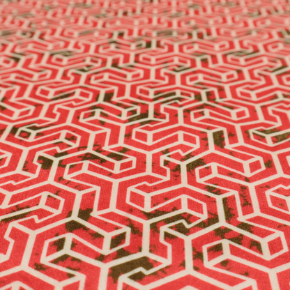 A coral red and off-white textile fabric featuring a small-scale, densely repeating interlocking geometric pattern. Fine off-white outlines construct complex, three-dimensional isometric block formations against a vibrant coral red background. Distressed, dark brown speckled textures are integrated throughout, creating a weathered, organic contrast to the sharp geometric structure.