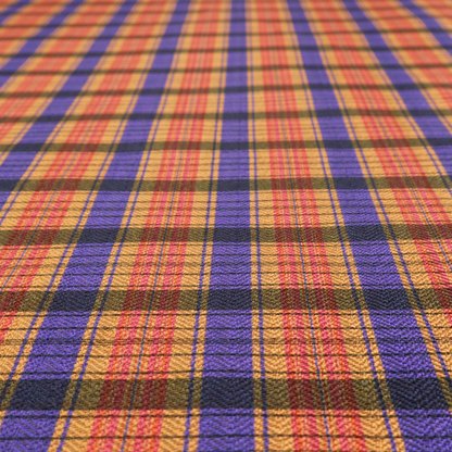A vibrant, multi-coloured plaid fabric featuring a complex grid of horizontal and vertical stripes. The pattern blends deep indigo, golden ochre, and olive green, highlighted by thin, high-contrast stripes of bright violet and coral pink.