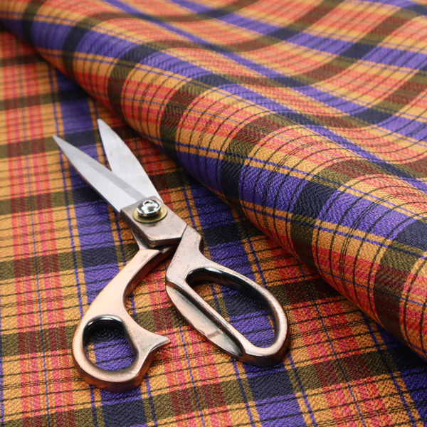 A vibrant, multi-coloured plaid fabric featuring a complex grid of horizontal and vertical stripes. The pattern blends deep indigo, golden ochre, and olive green, highlighted by thin, high-contrast stripes of bright violet and coral pink.