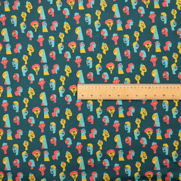 This fabric features a whimsical, repeating pattern on a dark teal background, decorated with stylized, colourful illustrations of diverse human faces in shades of bright yellow, light blue, and coral pink.