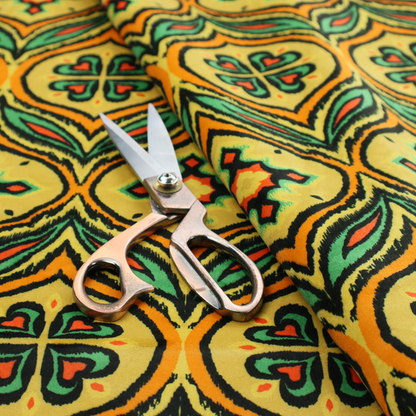 Fabric print featuring a repeating retro ikat ogee pattern. The geometric design displays a lattice of mustard yellow onion arches, green leaf shapes, and four-leaf clover medallions outlined in fuzzy black brushstrokes with pops of coral orange.