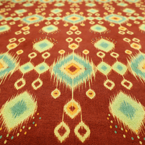Fabric print featuring a repeating vertical ikat pattern. The design showcases distinct diamond and teardrop motifs with feathered edges in soft sage green, teal, and mustard yellow over a solid terracotta red background.