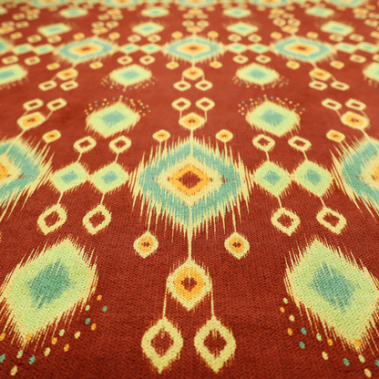 Fabric print featuring a repeating vertical ikat pattern. The design showcases distinct diamond and teardrop motifs with feathered edges in soft sage green, teal, and mustard yellow over a solid terracotta red background.