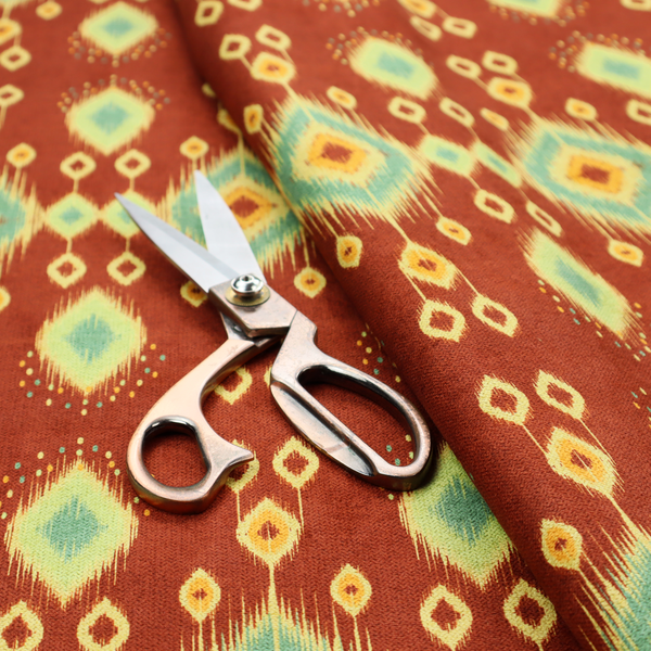 Fabric print featuring a repeating vertical ikat pattern. The design showcases distinct diamond and teardrop motifs with feathered edges in soft sage green, teal, and mustard yellow over a solid terracotta red background.