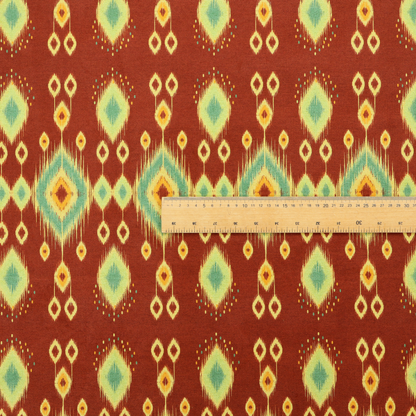 Fabric print featuring a repeating vertical ikat pattern. The design showcases distinct diamond and teardrop motifs with feathered edges in soft sage green, teal, and mustard yellow over a solid terracotta red background.