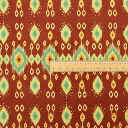 Fabric print featuring a repeating vertical ikat pattern. The design showcases distinct diamond and teardrop motifs with feathered edges in soft sage green, teal, and mustard yellow over a solid terracotta red background.