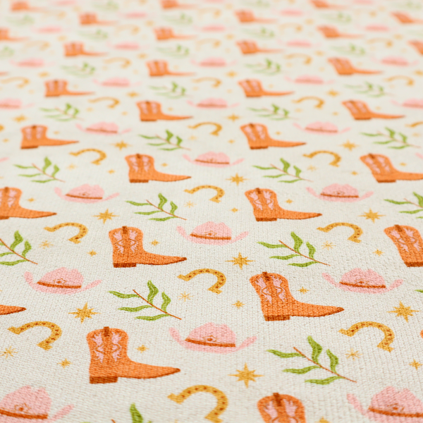 A repeating pattern of cowboy-themed illustrations on a cream-coloured fabric backdrop. The design features light orange cowboy boots, pale pink cowboy hats, golden horseshoes, small green leafy sprigs, and delicate four-pointed stars scattered throughout.