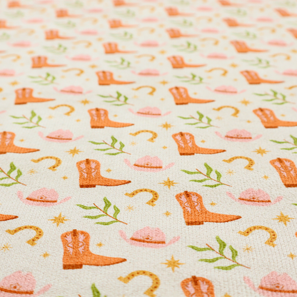 A repeating pattern of cowboy-themed illustrations on a cream-coloured fabric backdrop. The design features light orange cowboy boots, pale pink cowboy hats, golden horseshoes, small green leafy sprigs, and delicate four-pointed stars scattered throughout.