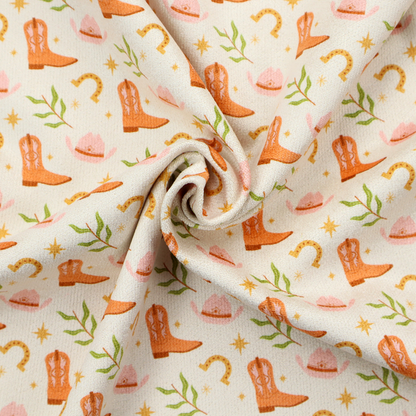 A repeating pattern of cowboy-themed illustrations on a cream-coloured fabric backdrop. The design features light orange cowboy boots, pale pink cowboy hats, golden horseshoes, small green leafy sprigs, and delicate four-pointed stars scattered throughout.