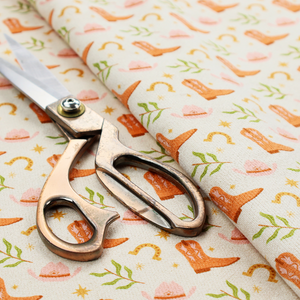 A repeating pattern of cowboy-themed illustrations on a cream-coloured fabric backdrop. The design features light orange cowboy boots, pale pink cowboy hats, golden horseshoes, small green leafy sprigs, and delicate four-pointed stars scattered throughout.