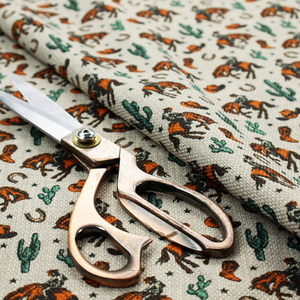 A seamless, repeating pattern featuring traditional western motifs in a classic comic or illustration style, set against a textured, light oatmeal or linen-look background. The colour palette relies heavily on bold terracotta orange, deep teal green, and dark brown accents.