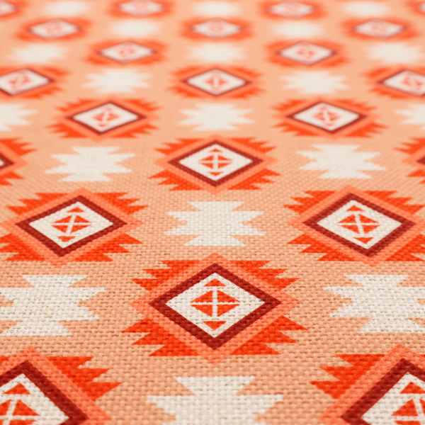 A uniform, repeating geometric pattern featuring southwestern and tribal-inspired motifs printed on a linen-textured background. The colour palette relies on warm, sun-baked tones, including a soft peach backdrop, vibrant terracotta orange, rust brown, and cream accents.