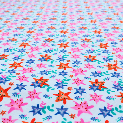 A dense, small-scale tropical ditsy pattern. The design consists of repeating star-shaped lily blossoms in bright shades of orange, magenta pink, and medium blue, accented with tiny teal green leafy sprigs and miniature blue flowers, all distributed across a solid light sky-blue background.