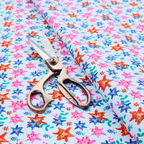 A dense, small-scale tropical ditsy pattern. The design consists of repeating star-shaped lily blossoms in bright shades of orange, magenta pink, and medium blue, accented with tiny teal green leafy sprigs and miniature blue flowers, all distributed across a solid light sky-blue background.