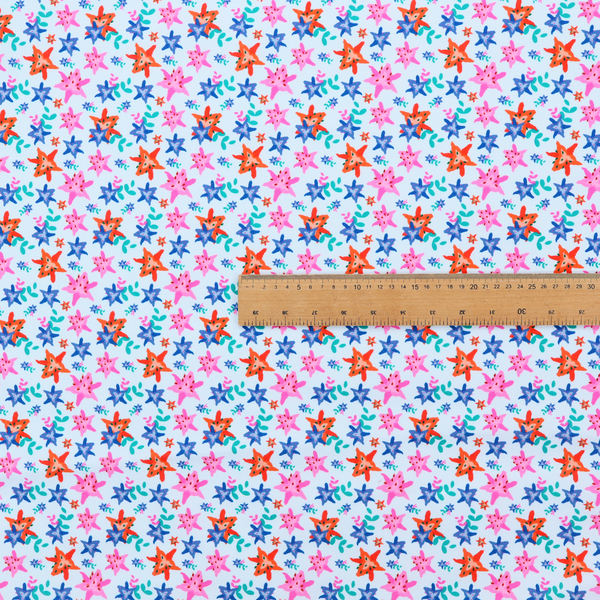 A dense, small-scale tropical ditsy pattern. The design consists of repeating star-shaped lily blossoms in bright shades of orange, magenta pink, and medium blue, accented with tiny teal green leafy sprigs and miniature blue flowers, all distributed across a solid light sky-blue background.
