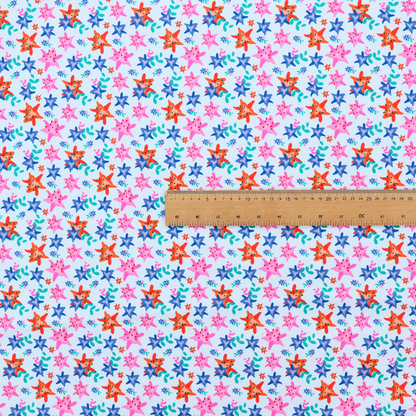 A dense, small-scale tropical ditsy pattern. The design consists of repeating star-shaped lily blossoms in bright shades of orange, magenta pink, and medium blue, accented with tiny teal green leafy sprigs and miniature blue flowers, all distributed across a solid light sky-blue background.