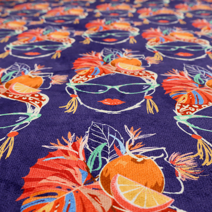A repeating pattern on dark purple fabric featuring stylized line-art faces of women wearing light green cat-eye sunglasses, bright red lipstick, and elaborate tropical headwraps adorned with orange slices and palm leaves.