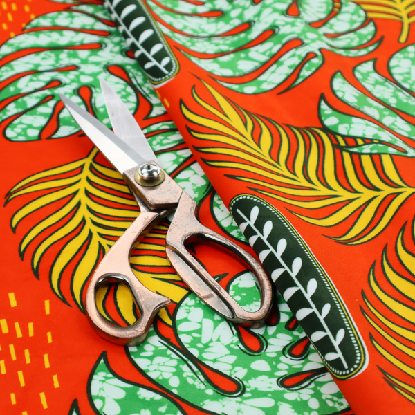 Fabric featuring a vibrant, repeating tropical pattern with textured green monstera leaves, yellow ferns, and dark green patterned seed pods on a bright orange background.