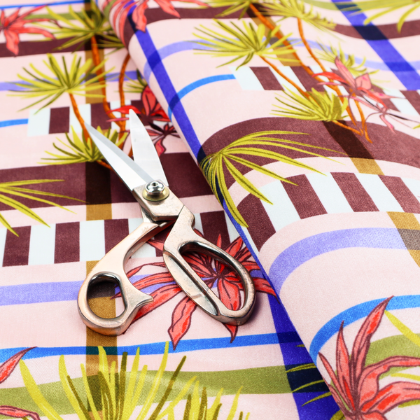 An eclectic, geometric tropical fabric pattern featuring a unique overlay design. The background is a structured windowpane plaid layout composed of intersecting lines in cobalt blue, olive green, and chocolate brown across a dusty pink base. Layered over this grid is a repeating botanical print of tropical plants, featuring fan palms in lime green and slender fronds in terracotta pink.