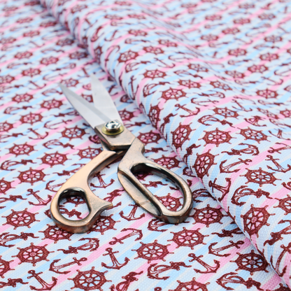 A nautical-themed fabric pattern featuring diagonal stripes in light blue and pink, scattered with dark red ship's wheels and anchors.