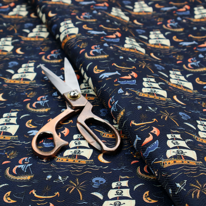 A dark navy blue fabric patterned with a repeating pirate-themed design featuring multi-sailed galleon ships with skull-and-crossbones flags sailing on stylized waves, accompanied by smaller elements including blue whales, crescent moons, skulls, tropical palm trees, and anchors.
